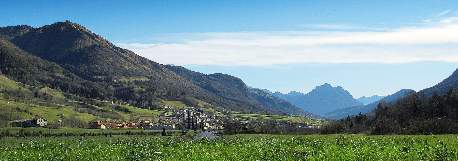 Cerete Basso con il campanile della parrocchiale tra i prati verdi dell'altopiano, con le montagne della bergamasca sullo sfondo