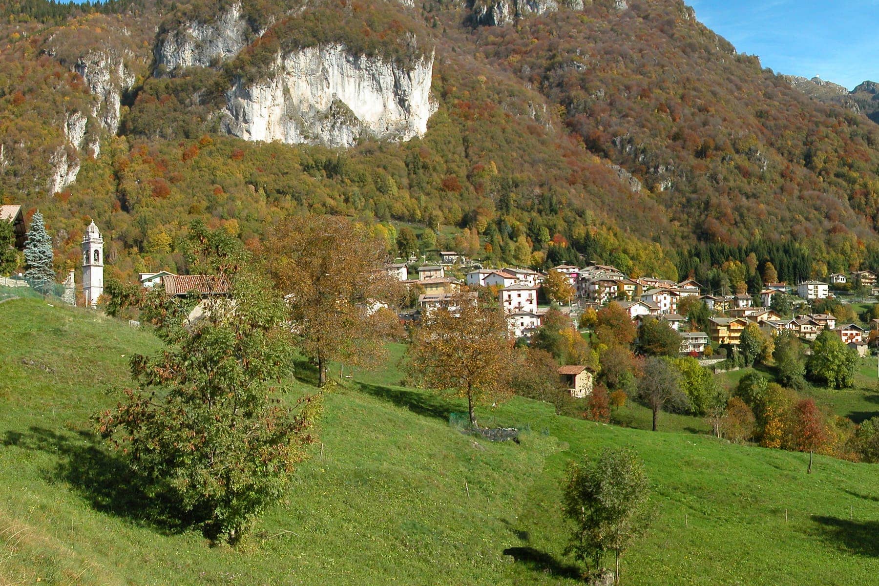 Cornalba in autunno con il campanile, le case sui prati e la parete bianca della falesia sopra i boschi