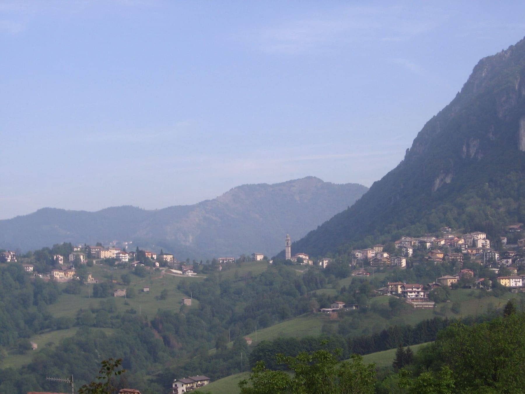 Cornalba con il campanile e le case sui prati sotto le pareti calcaree, in una mattina di foschia primaverile