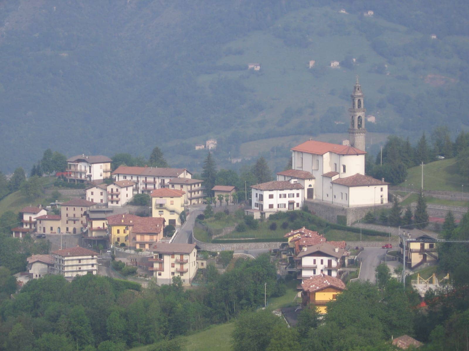 Il centro di Costa Serina con la parrocchiale dei Santi Lorenzo e Ambrogio e il campanile, tra i boschi