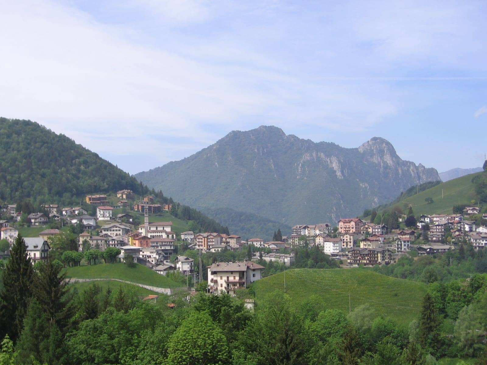 Il paese di Oltre il Colle in primavera, con il campanile della parrocchiale tra le case e i monti boscosi alle spalle
