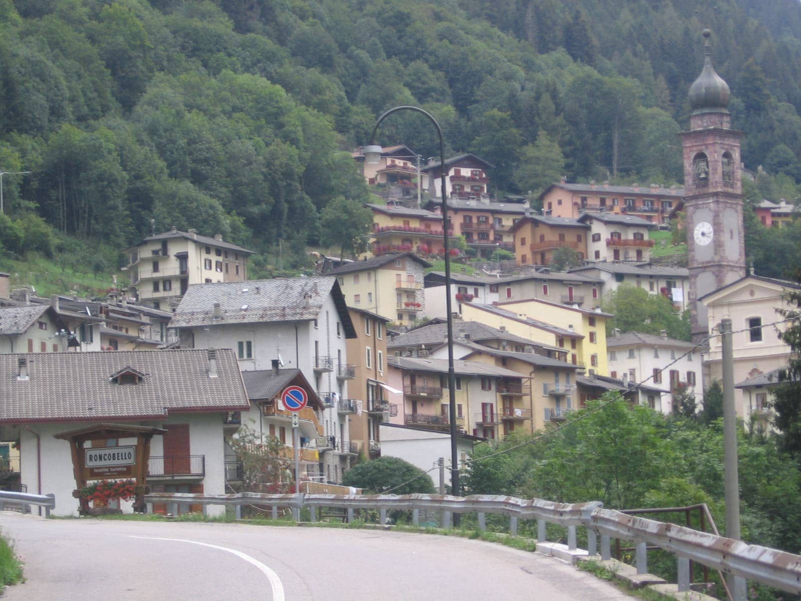 Il centro di Roncobello dalla strada d'ingresso, con le case colorate e il campanile della parrocchiale tra i boschi dell'alta Valle Brembana