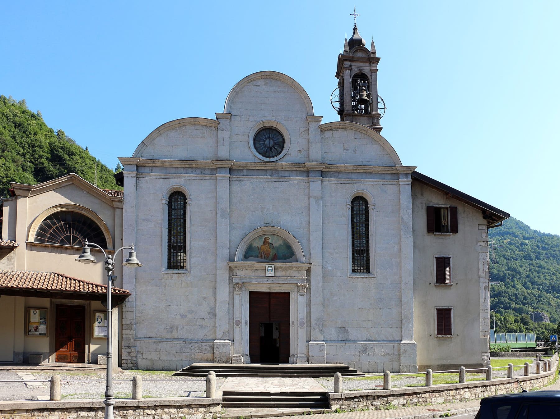 Facciata in pietra chiara della chiesa parrocchiale di San Giacomo Maggiore a Sedrina, con il campanile e il versante boscoso alle spalle