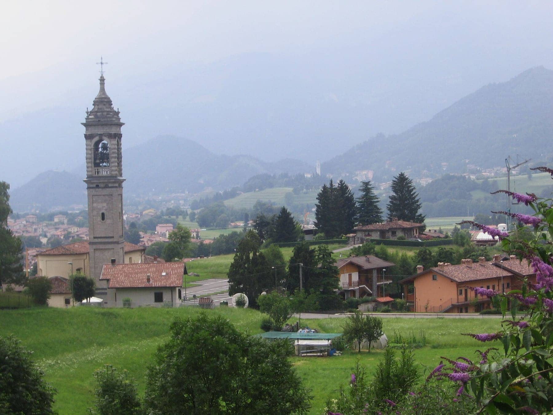 Songavazzo con il campanile della chiesa di San Bartolomeo e i prati verdi, sullo sfondo Clusone e i monti dell'altopiano
