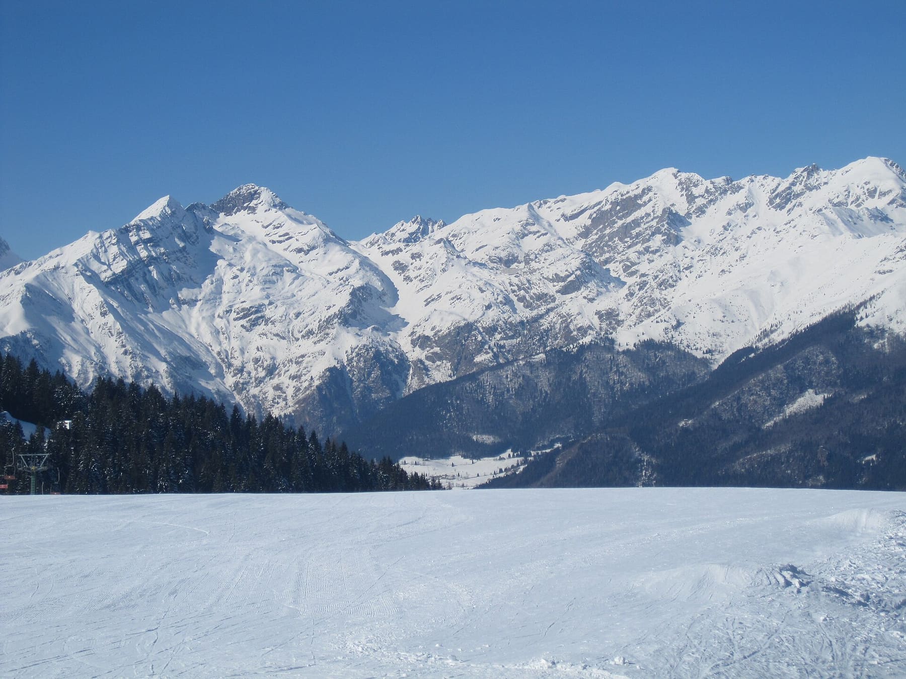 Pista da sci battuta di fresco sotto un cielo terso, con una seggiovia tra gli abeti e le cime innevate delle Orobie bergamasche